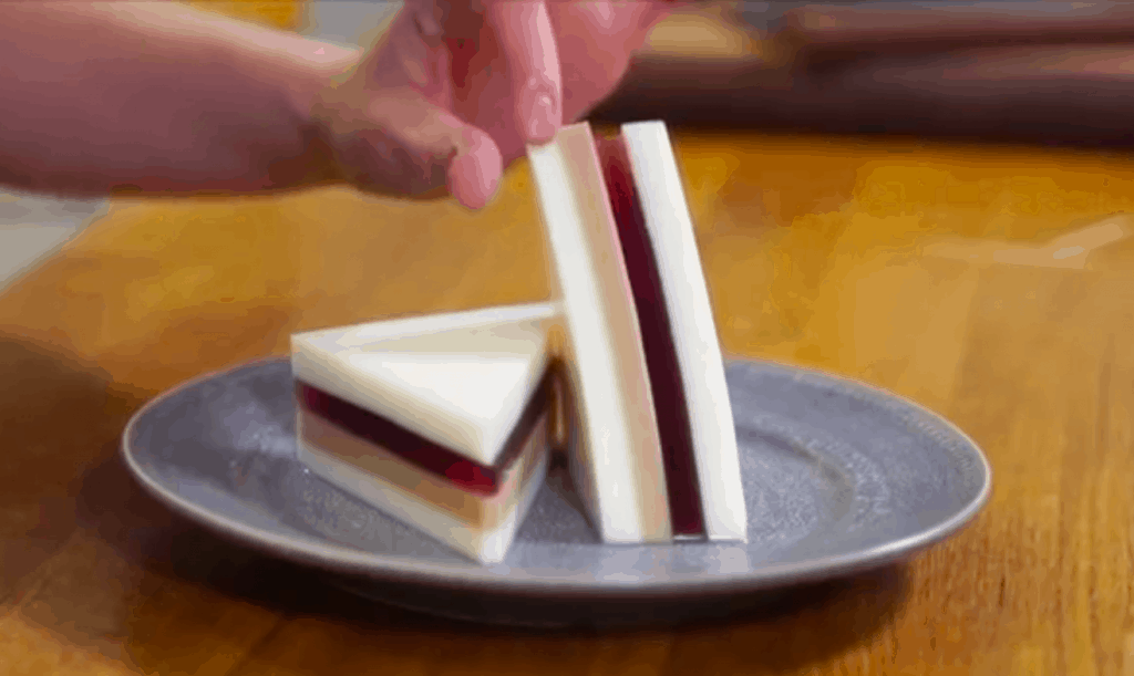 Crustless peanut butter and jelly sandwich cut on the bias, one half flat on plate, the other half resting against it, rising vertically. Sandwich has a glossy, almost plastic apperance.