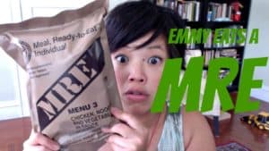 Emmy holding up an MRE with a surprised expression. Green text saying "Emmy Tastes a MRE."