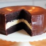 A two-layered chocolate cake sitting in a cake stand with a thick white filling coated in a chocolate glaze.