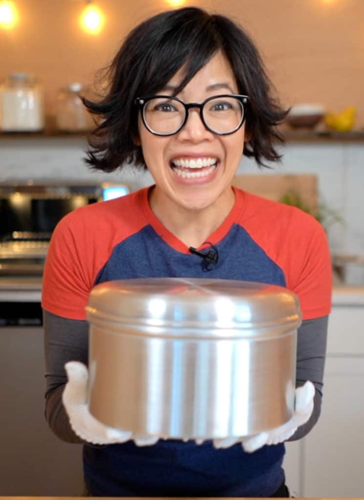 A bespectacled woman with short black hair holding a metal jachnun tin.