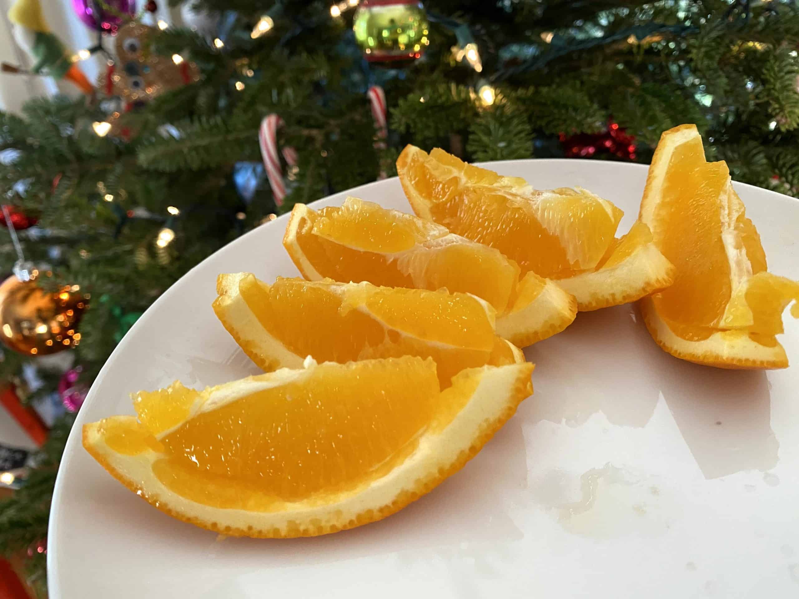 Sliced orange wedges on a white plate with a Christmas tree in the background. 