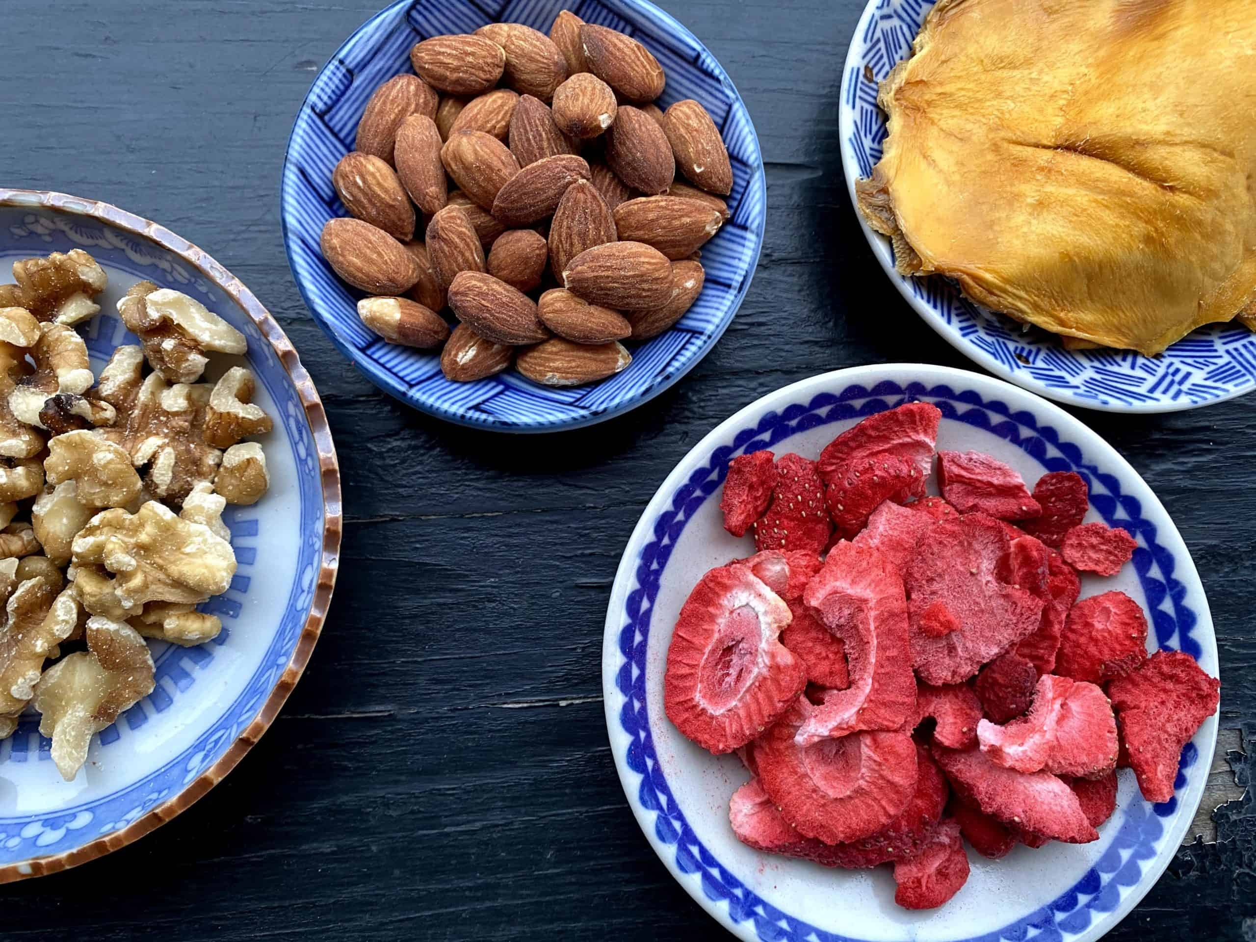 A variety of saucers of healthy snacks including almonds,walnuts,freeze-dried strawberries, and dried mango on a black background.