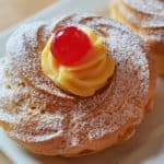 A cream puff topped with pastry cream and a maraschino cherry dusted with powdered sugar on a white plate.