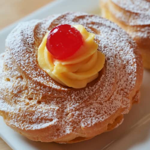 A cream puff topped with pastry cream and a maraschino cherry dusted with powdered sugar on a white plate.