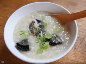 White bowl with rice porridge, wooden spoon, chopped onions and century eggs.