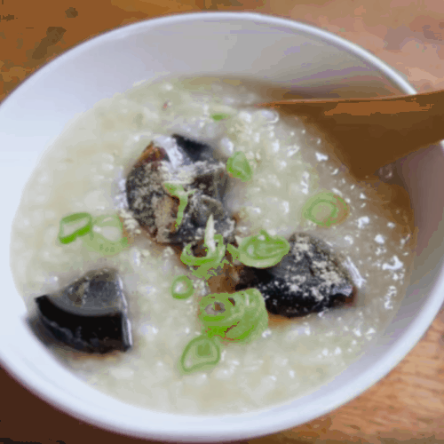 White bowl with rice porridge, wooden spoon, chopped onions and century eggs.