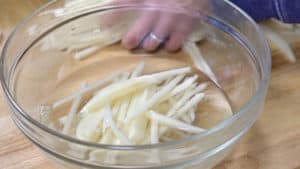 Clear glass bowl with uncooked French fries resting in sugar-srup water.