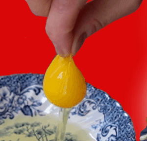 Thumb and forefinger pinching and holding up egg yolk with white dripping down into dish.