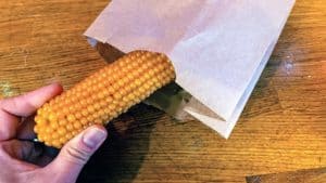Thumb and forefinger putting unpopped corn-on-the-cob into brown paper bag.