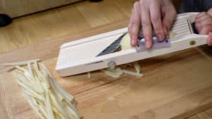 Uncooked French fries laying on cutting board beneath mandoline slicing potato.