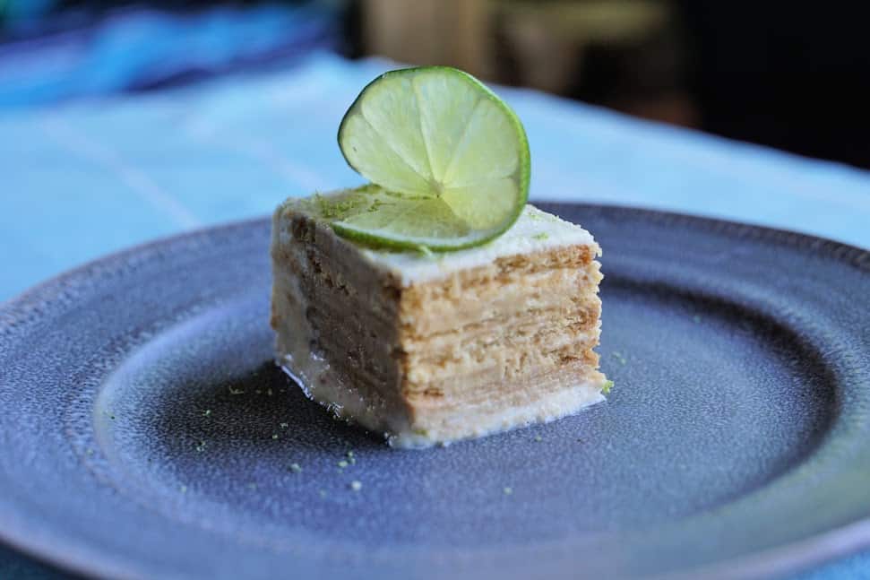 Square of layered Maria biscuits cake with frosting and wedge of lime on plate.