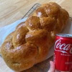 Hand holding small sized Coca-Cola next to a braided loaf of freshly backed bread.