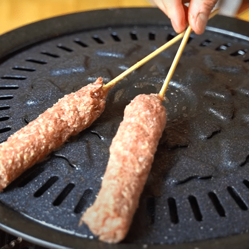 A hand holding two skewered kabobs over an indoor grill top.